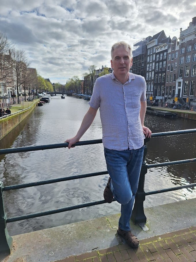 Justin Smith in Amsterdam with a canal and typical dutch brick buildings in the background with a cloudy sky.