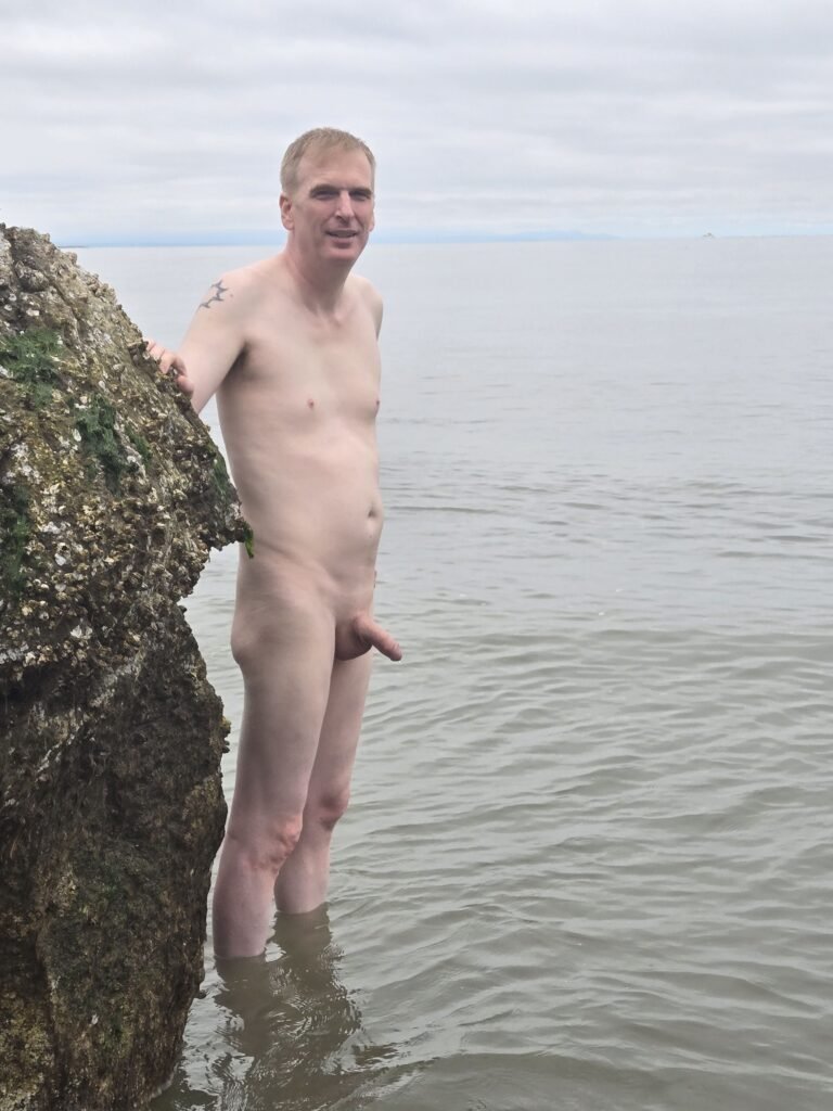 Naked man standing in the ocean in ankle deep water next to a rock on a nude beach in Vancouver.