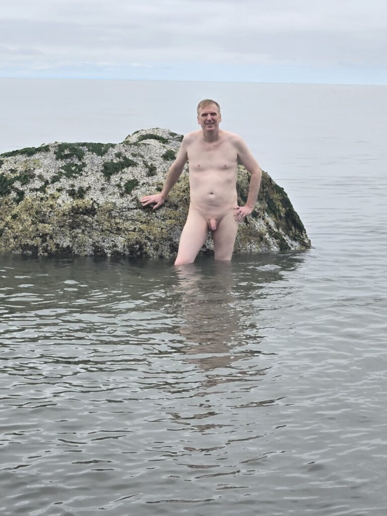 Naked man standing in the ocean in Vancouver in knee deep water posing next to a large partially submerged rock.