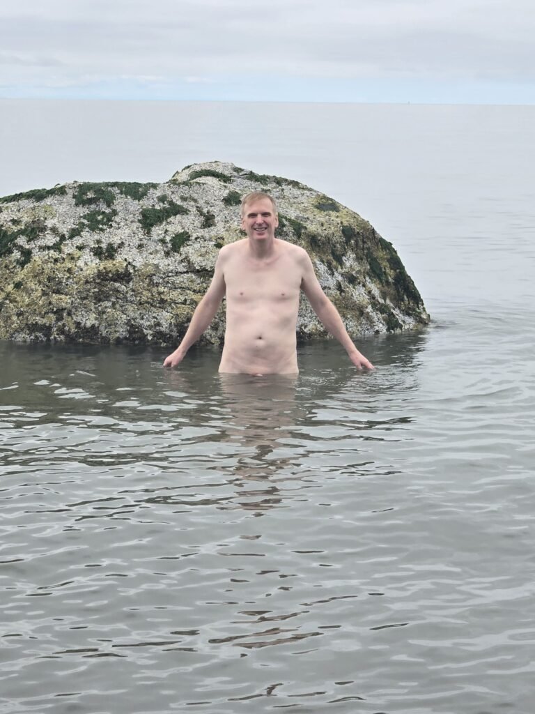 naked man in the ocean posing next to a large rock.