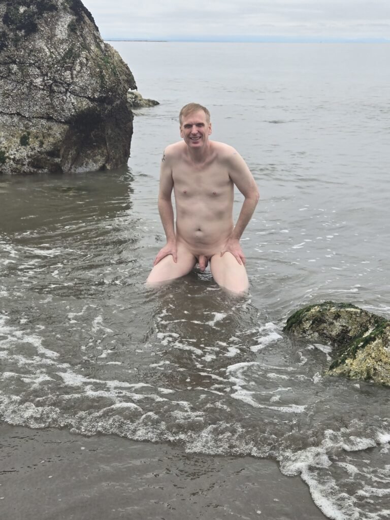 Naked man kneeling in the water at Wreck Beach in Vancouver on a cloudy day.