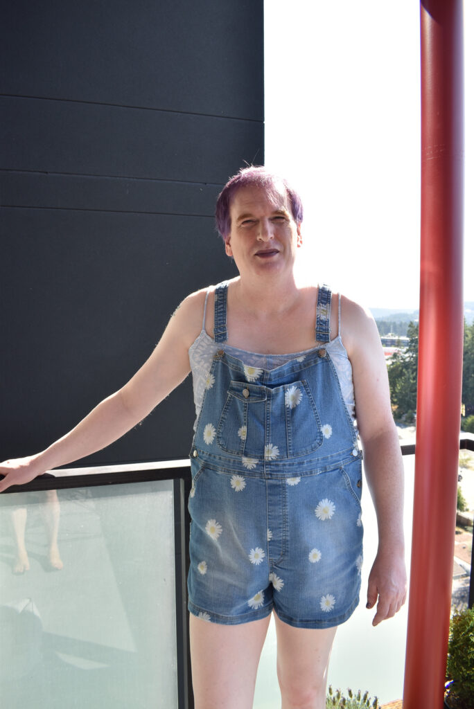male cross dresser wearing denim overalls with white flowers standing outside.